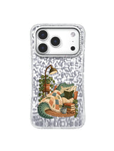 Muted teal crocodile reading under a patchwork blanket on an iPhone 17 Pro case illustration.