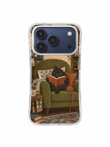 Charcoal cat reading between patterned cushions with yarn beside the olive armchair on an iPhone 17 Pro case