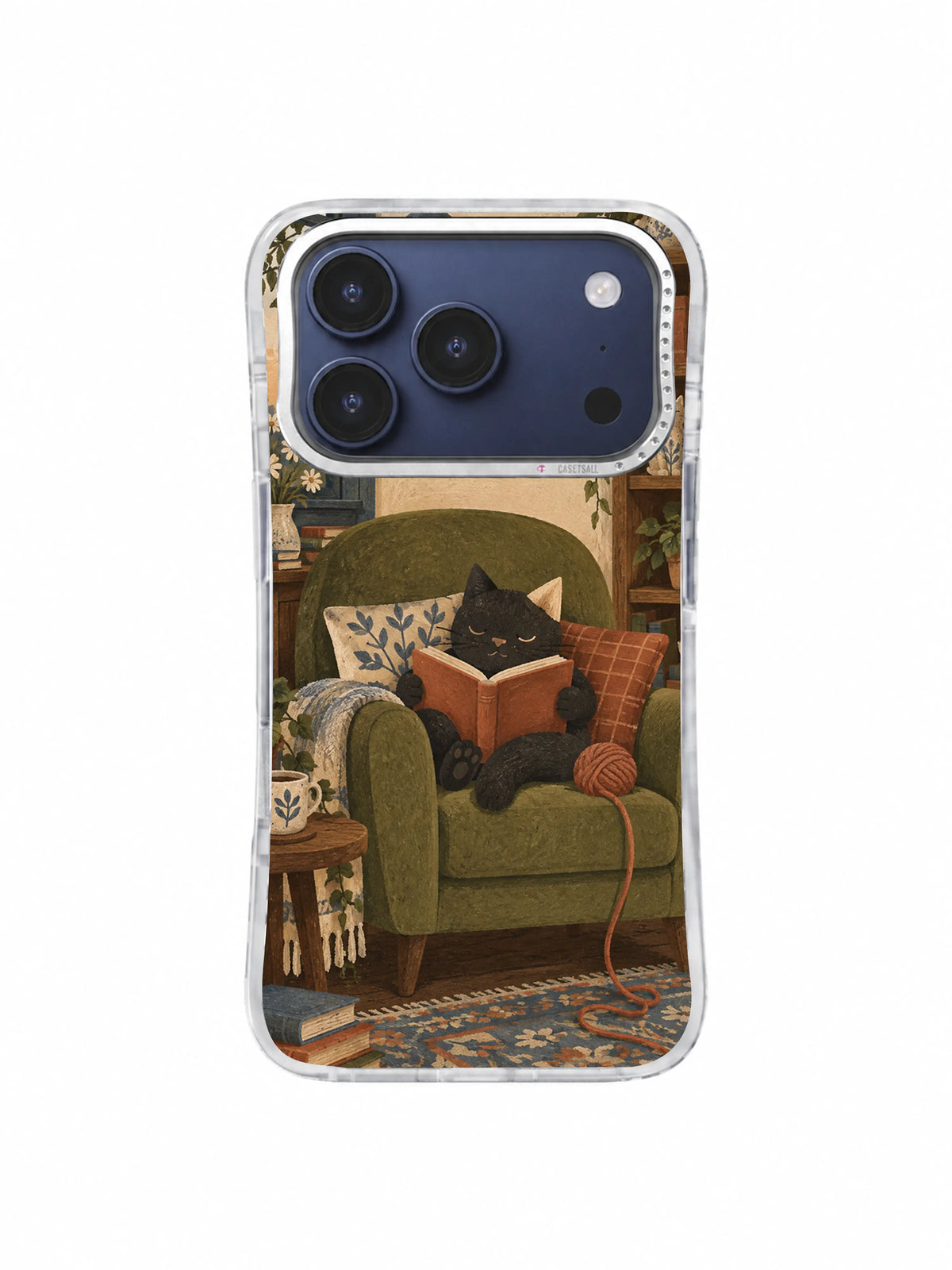 Charcoal cat reading between patterned cushions with yarn beside the olive armchair on an iPhone 17 Pro case
