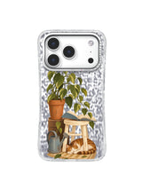 Terracotta plant leaning over an open book while a striped cat rests beneath the stool on an iPhone 17 Pro case