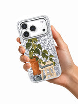 Hand holding a clear iPhone 17 Pro case decorated with plant, book, stool, and watering can art