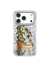 Green leaves arching toward an open dusty-blue book beside a watering can on an iPhone 17 Pro case