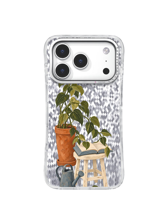 Tall terracotta potted plant bending leafy stems toward an open blue book on a cream stool on an iPhone 17 Pro case