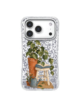 Tall indoor plant reading over a book on a pale stool with warm terracotta and olive tones on an iPhone 17 Pro case