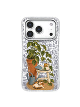 Leafy potted plant, open book, watering can, and resting cat in a quiet reading corner on an iPhone 17 Pro case