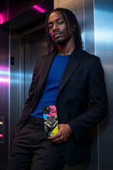 Adult man in a neon-lit elevator lobby holding the Rose Cobra Voltage iPhone 15 Plus case with Pink Bloom Strike artwork