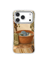 Tiny whale reading in a terracotta washbasin with an olive towel and cream cup on an iPhone 17 Pro case