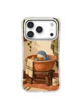 Tiny blue whale in a terracotta washbasin on a wooden stool with an olive towel on an iPhone 17 Pro case